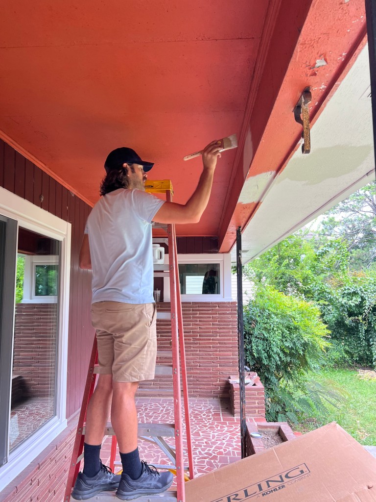 Photo of a man standing on a ladder painting a front porch. Porch contains custom mosaic brick tile flooring.